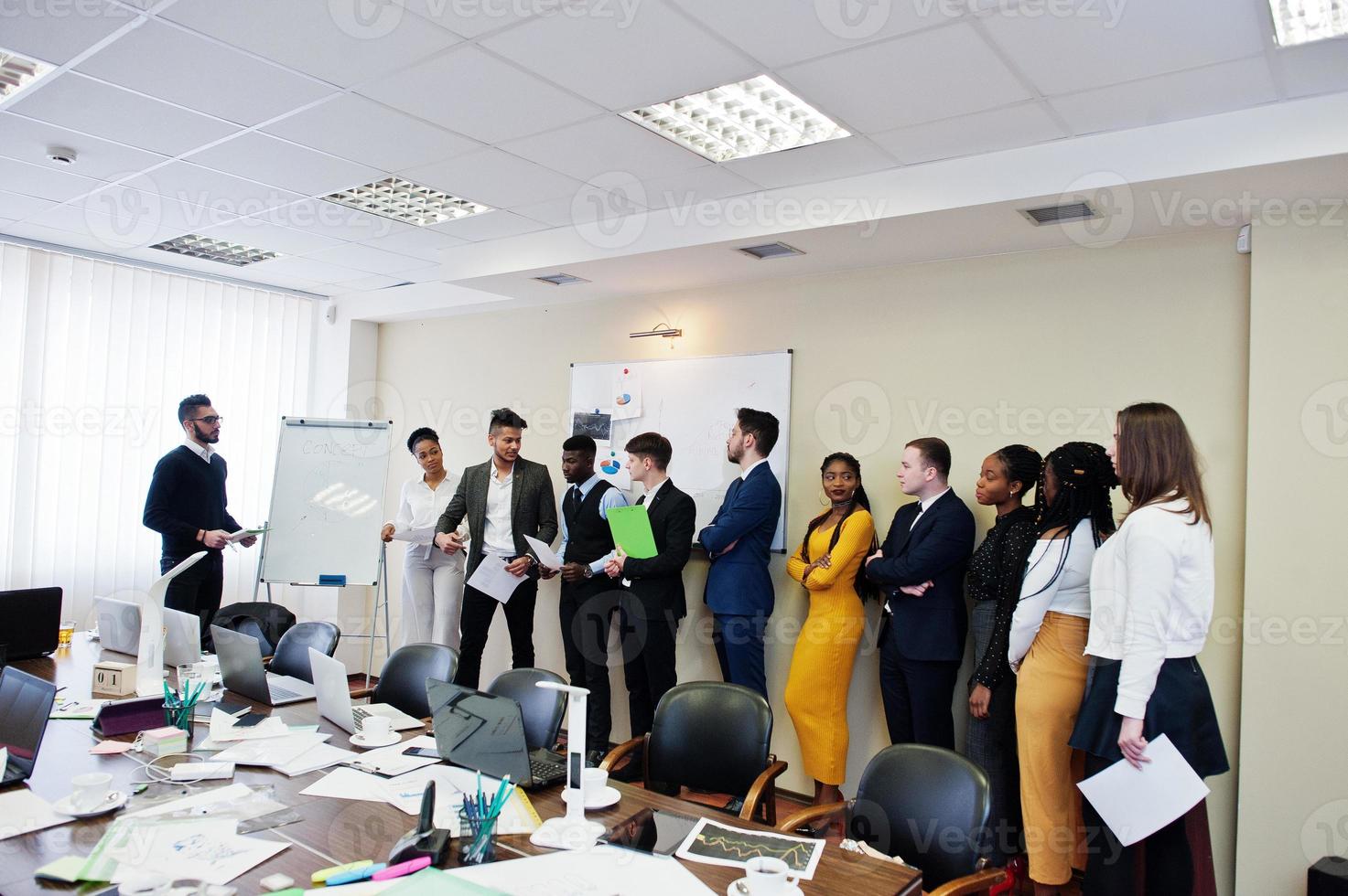 Mixed race business coach presenting report standing near whiteboard pointing on sales statistic shown on diagram and chart teach diverse company members gathered together in conference room. photo