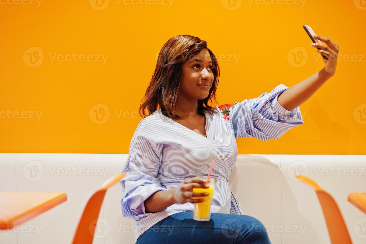 African woman sitting on cafe against orange wall with pineapple juice in hands and making selfie. photo