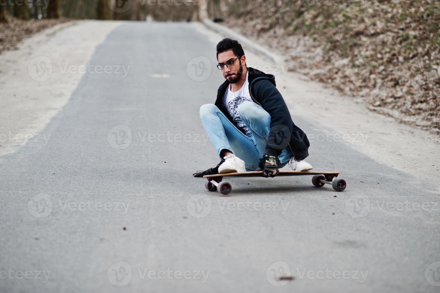 hombre árabe de estilo callejero en anteojos con longboard longboarding por el camino. foto