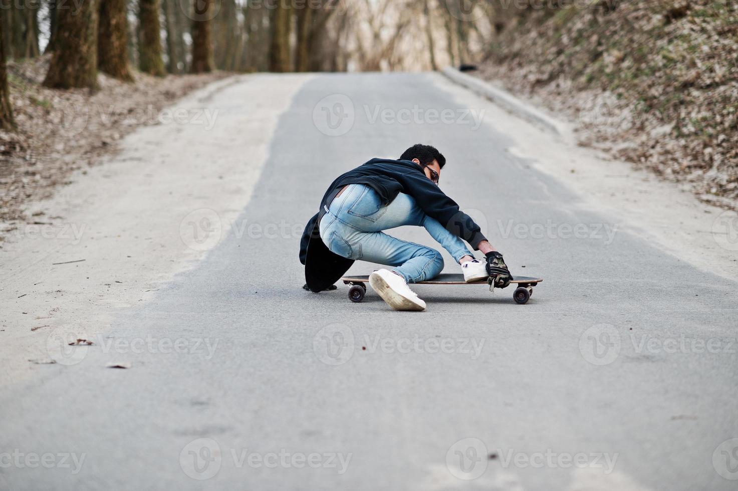 fallar al caer de una patineta. hombre árabe de estilo callejero en anteojos con longboard longboarding por el camino. foto