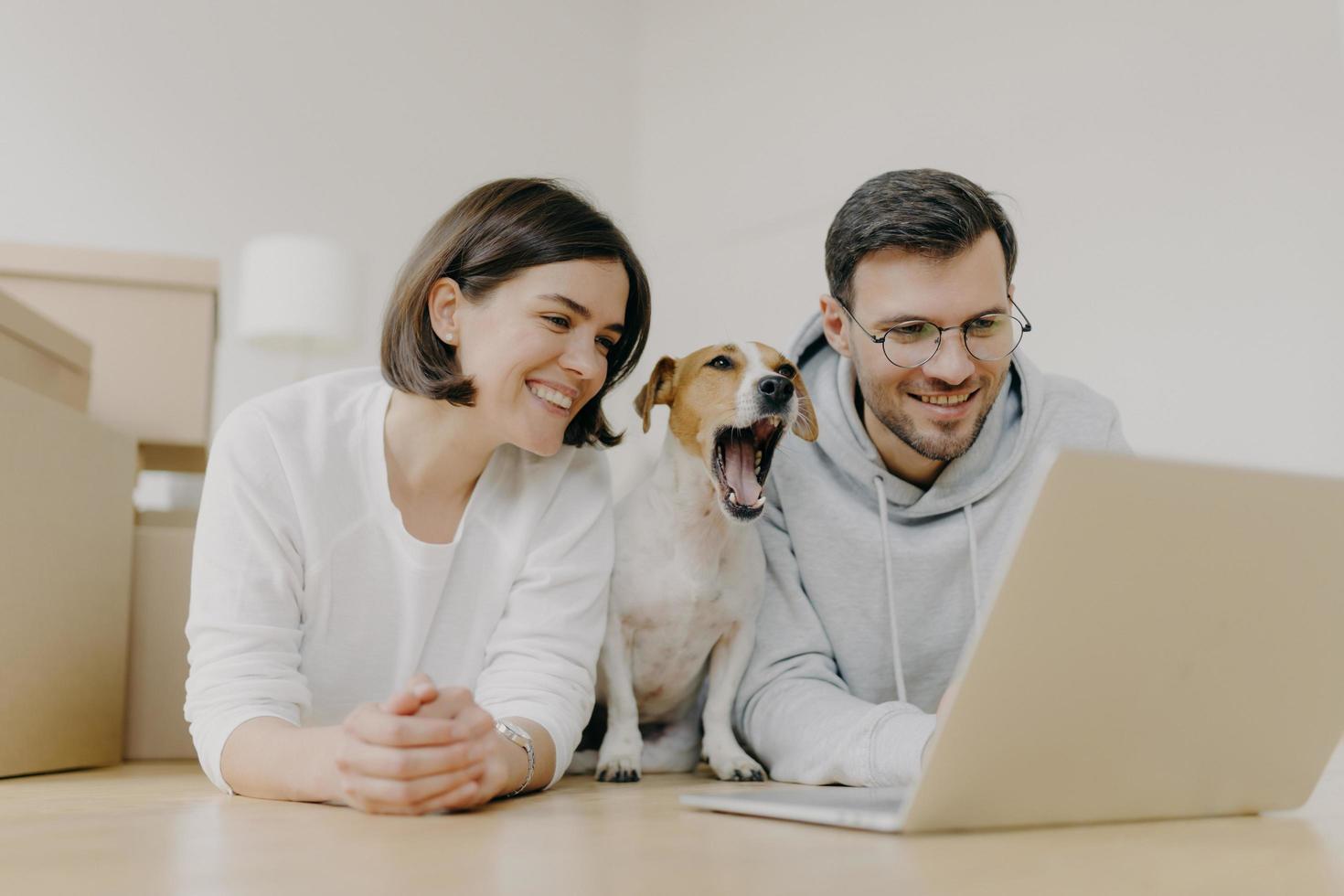 Photo of smiling woman and man work on modern laptop computer, dog yawns, buy furniture for new apartment, lie on floor in spacious light room, have glad expressions. Housewarming and repair concept