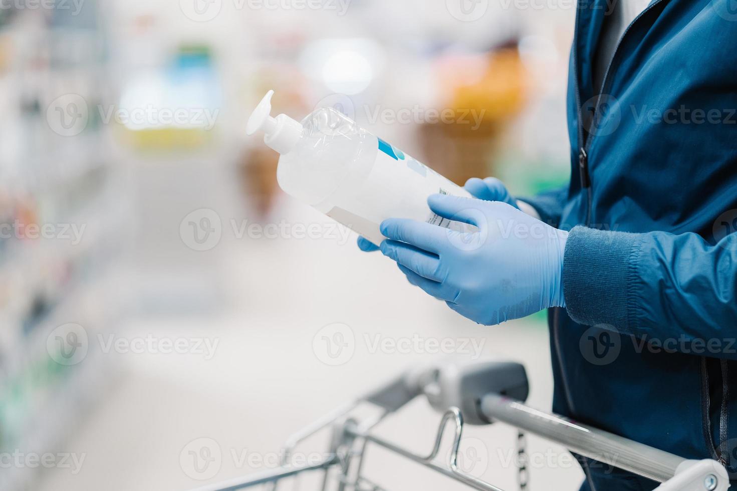 Horizontal shot of faceless man wears medical protective gloves during coronavirus outbreak, holds bottle of soap, cares about hygiene and disinfection during pandemic, makes shopping in store photo