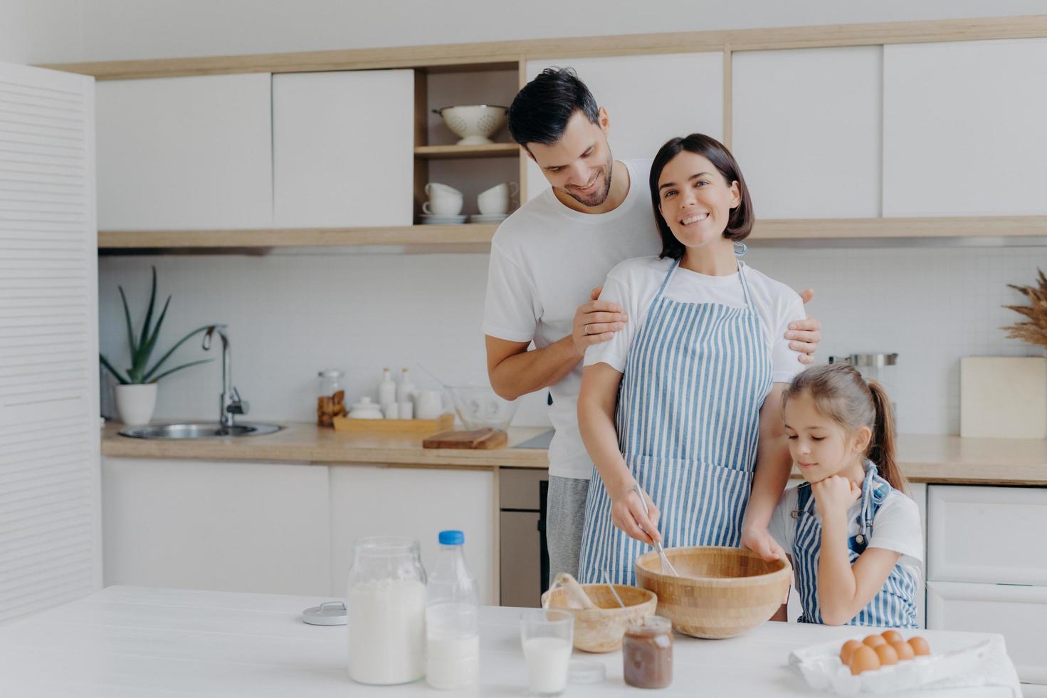 Happy family cook together at kitchen. Father, mother and dauther busy preparing delicious meal at home. Husband embraces wife who whisks and prepares dough, bake cookies. Food, togetherness photo