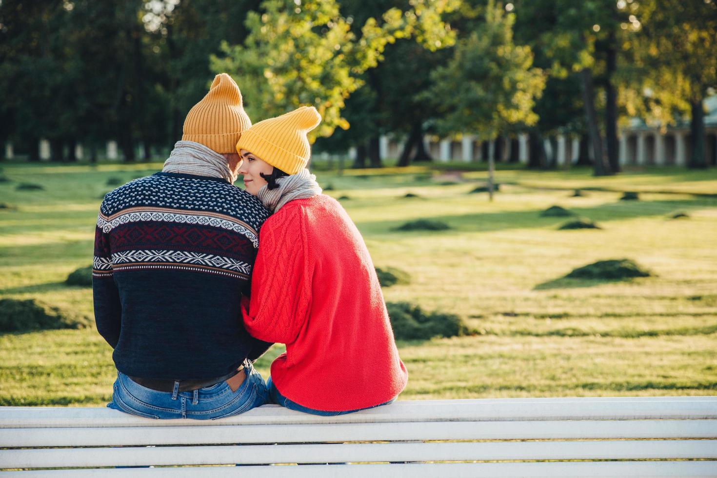 Lovely couple sit on bench together, wear warm clothes and knitted hats, embrace each other, express love and good relationship, enjoy beautiful landscapes and fresh air in park. Relationship concept photo