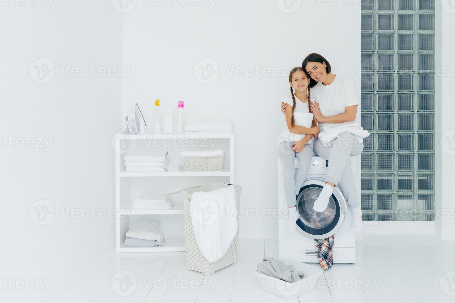 Shot of beautiful woman and his small daughter embrace and smile pleasantly, sit on washing machine, wash linen in laundry room, have friendly relationship, do laundry at home. Housework concept photo