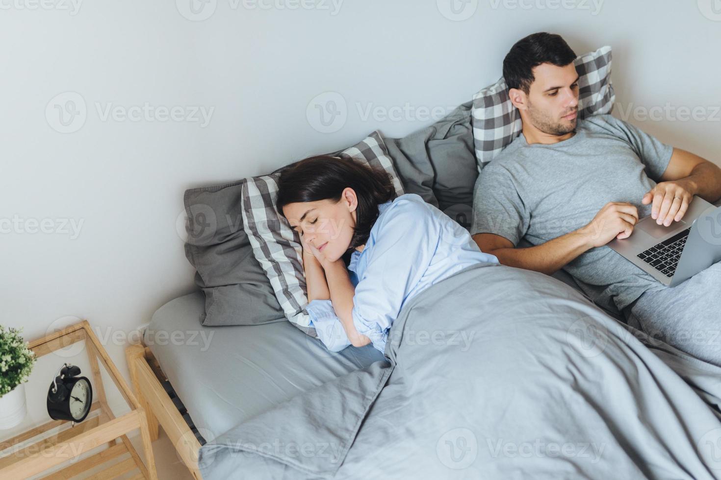 Pretty female sleeps in bed, sees pleasant dreams, while her husband works on laptop computer, prepares report, tries not to interrupt his wife sleep. Family couple in bedroom. Rest concept. photo
