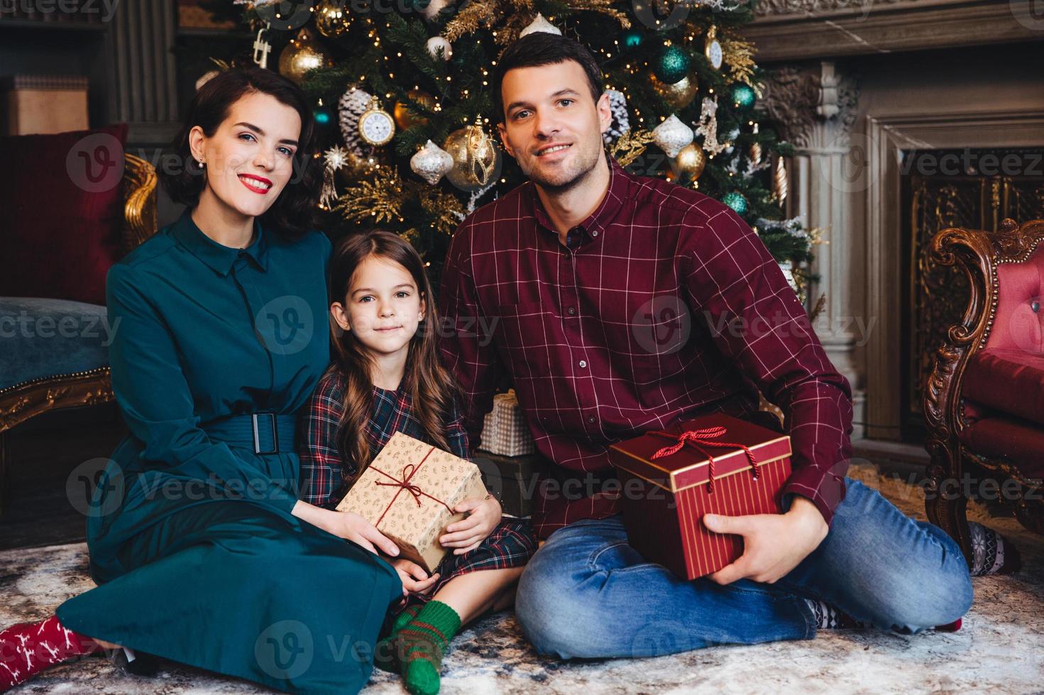 Father, mother and daughter enjoy holidays together, smile happily as sit in living room near decorated New Year tree, hold present wrapped in boxes, look delightfully into camera. Family concept photo