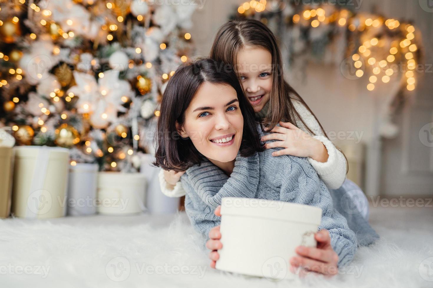 This present is for you Happy small kid embraces her affectionate mother who holds wrapped present, stands against decorated background with garlands and New Year tree. Domestic atmosphere. photo