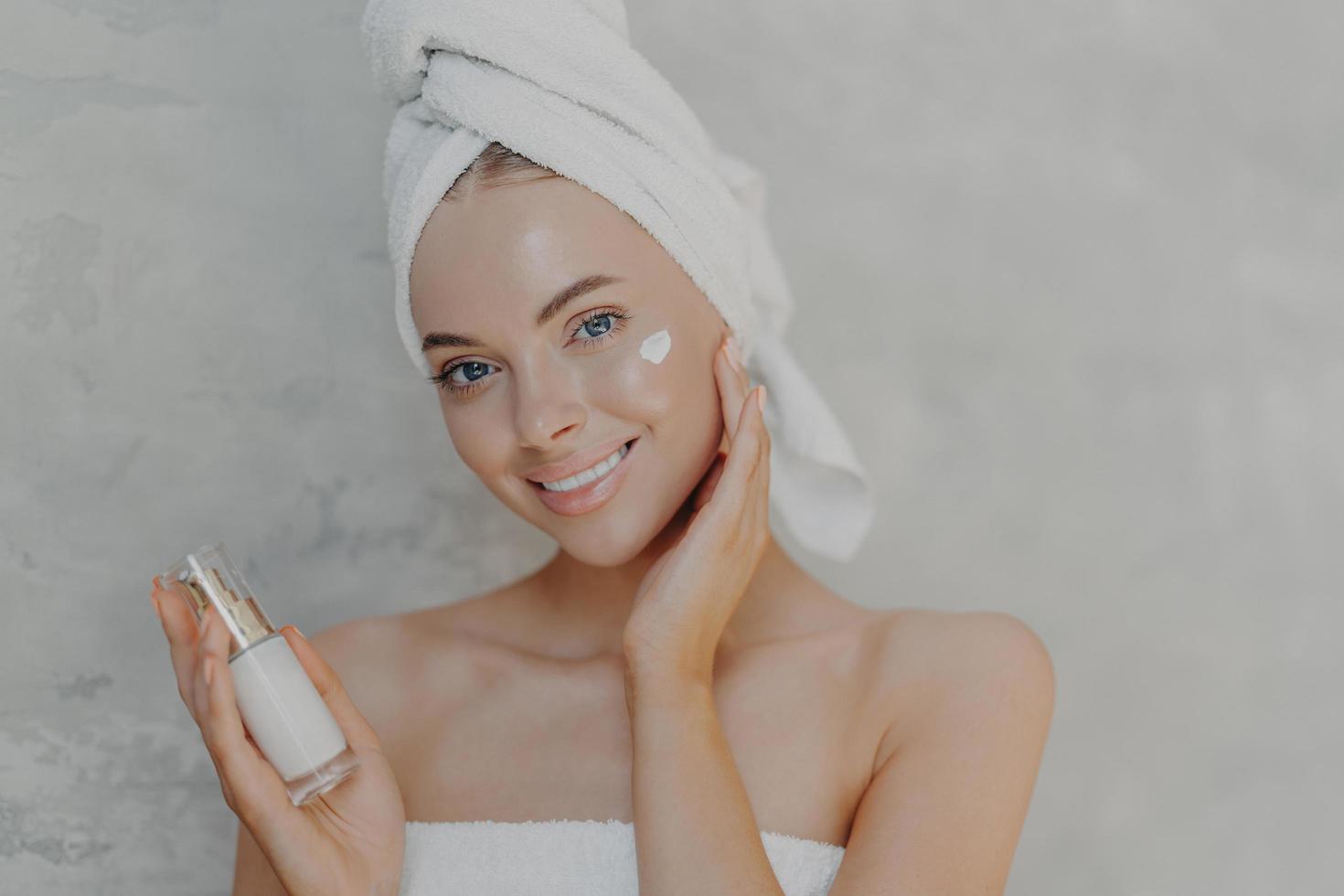 Close up shot of pretty young European woman with minimal makeup, applies moisturising cream on face, smiles gently and looks at camera, wears bath towel on head, isolated over grey background photo