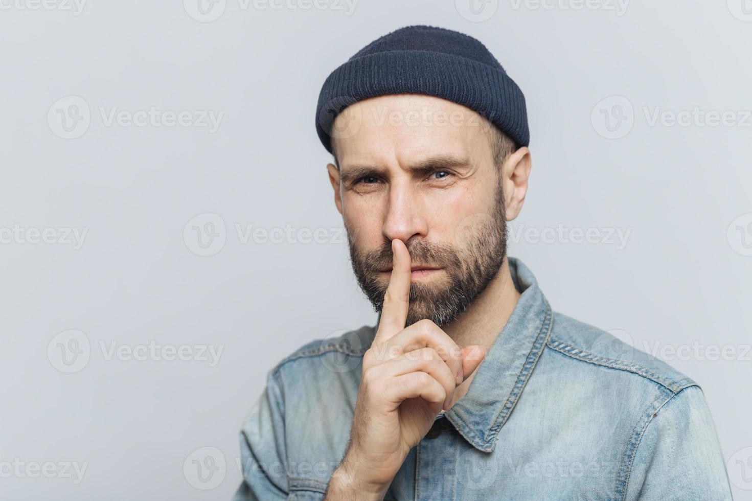 Serious pleasant looking bearded male with mysterious expression keeps fore finger on lips, asks to keep personal information confidential, wears stylish hat and denim shirt, isolated on grey wall photo