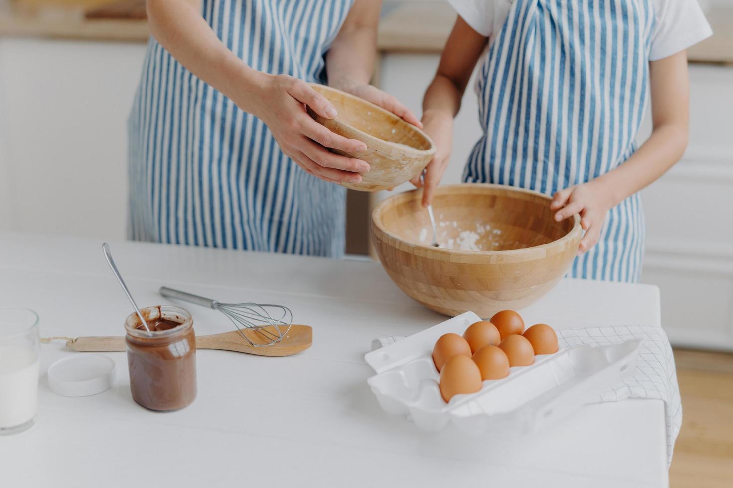 Cropped image of mothers and daughters hands mixing ingredients to prepare dough and bake tasty pastry, stand near kitchen table with eggs, melted chocolate in glass, wear striped blue aprons photo