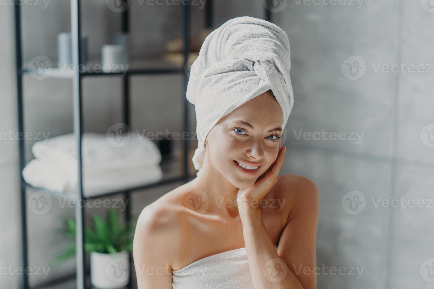 hermosa mujer europea con maquillaje toca la piel, tiene un maquillaje mínimo, tiene una piel saludable y brillante, envuelta en una toalla de baño, disfruta del descanso en casa. mujer spa posa en el baño. belleza, concepto de bienestar foto