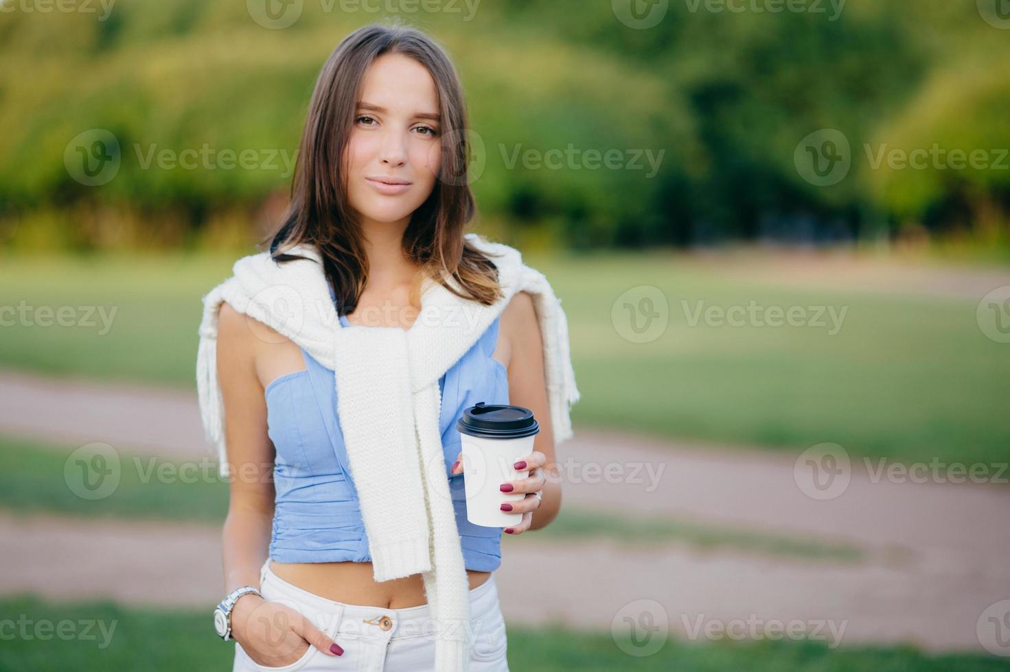 Outdoor shot of brunette woman dressed in casual t shirt, white trousers, keeps hand in pocket, drinks aromatic coffee during morning walk, stands against green nature background. Leisure concept photo