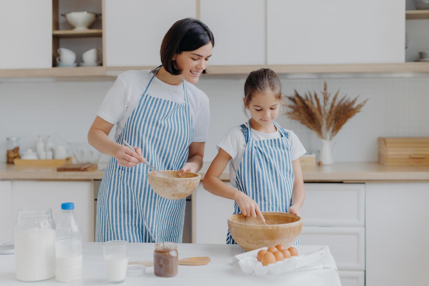 Preparing delicious meal and cooking together concept. Pleased mother looks attentively at how child whisks ingredients in bowl, wear aprons, stand in kitchen. Kid takes cooking lesson in mum photo