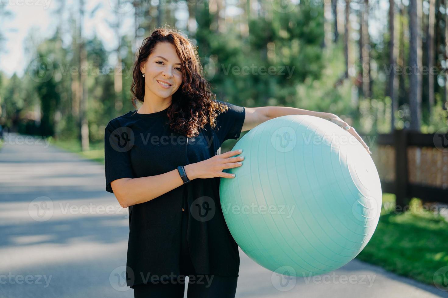 Fitness, sport and healthy lifestyle concept. Smiling curly brunette woman in black t shirt holds big fitness ball poses outside against forest background, breathes fresh air during sunny day photo