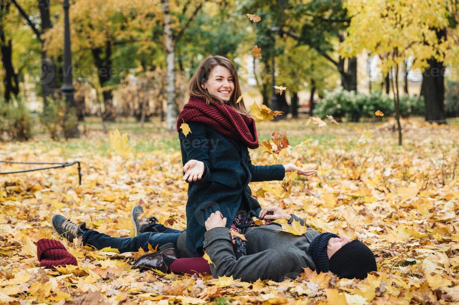 Positive woman wears knitted scarf, sits on boyfriend, throws yellow leaves in air, has good mood as enjoys last warm autumn days. Romantic couple spend weekends together, have fun. Love concept photo