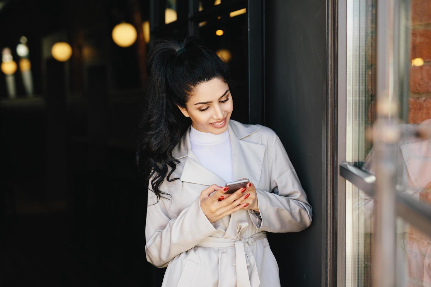 retrato de una mujer morena de moda con una apariencia agradable que tiene una bonita manicura con un impermeable blanco que sostiene un teléfono inteligente en sus manos con una sonrisa agradable que se alegra de recibir el mensaje foto