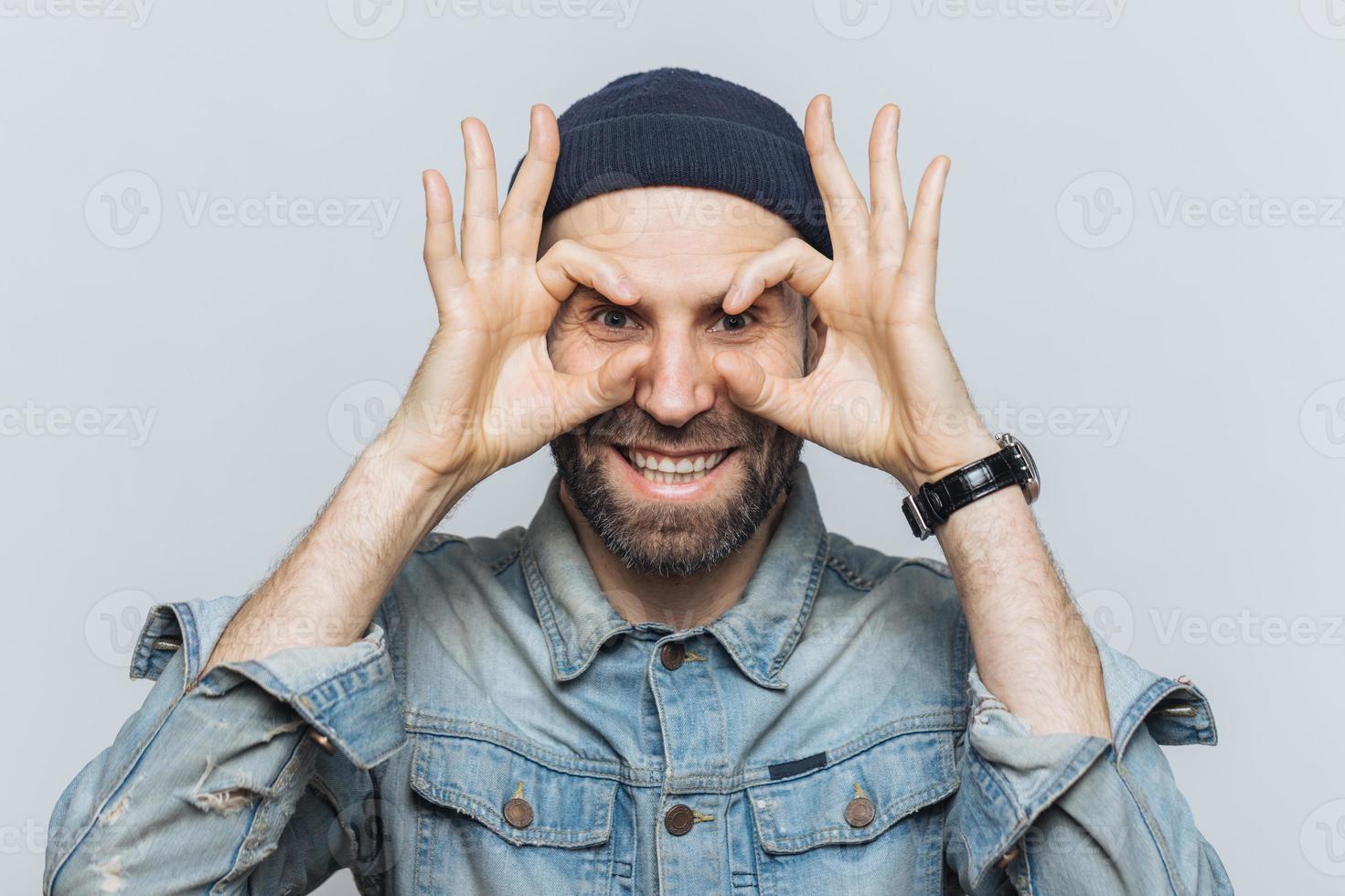Portrait of hppy bearded male makes eyewear with fingers, has stubble, wears jeans cloting, stands against white background. Funny middle aged man foolishes indoor, pretends wearing glasses. photo