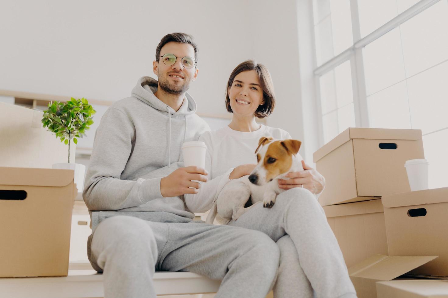 Shot of cheerful husband and wife pose together with pedigree dog, pose in empty room with no furniture, carton boxes with personal belongins, drink takeaway coffee, enjoy togetherness. Moving photo