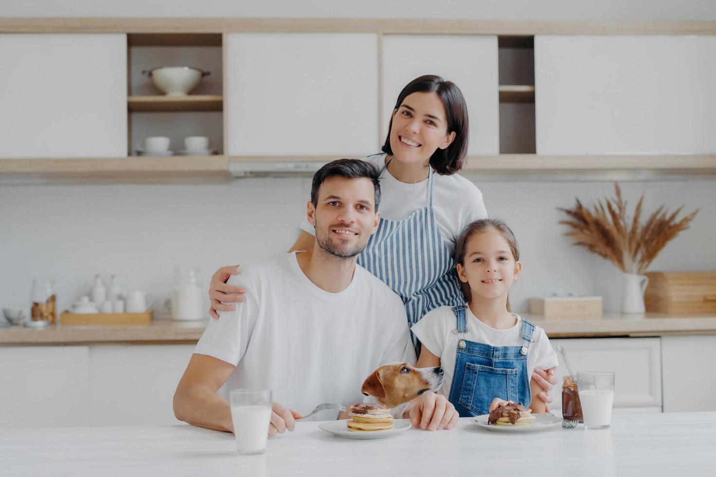 Smiling lovely mother and wife embrces with love daughter and husband, prepared delicious breakfast for them. Little child and father, their pedigree dog eat pancakes in kitchen. Friendly family photo