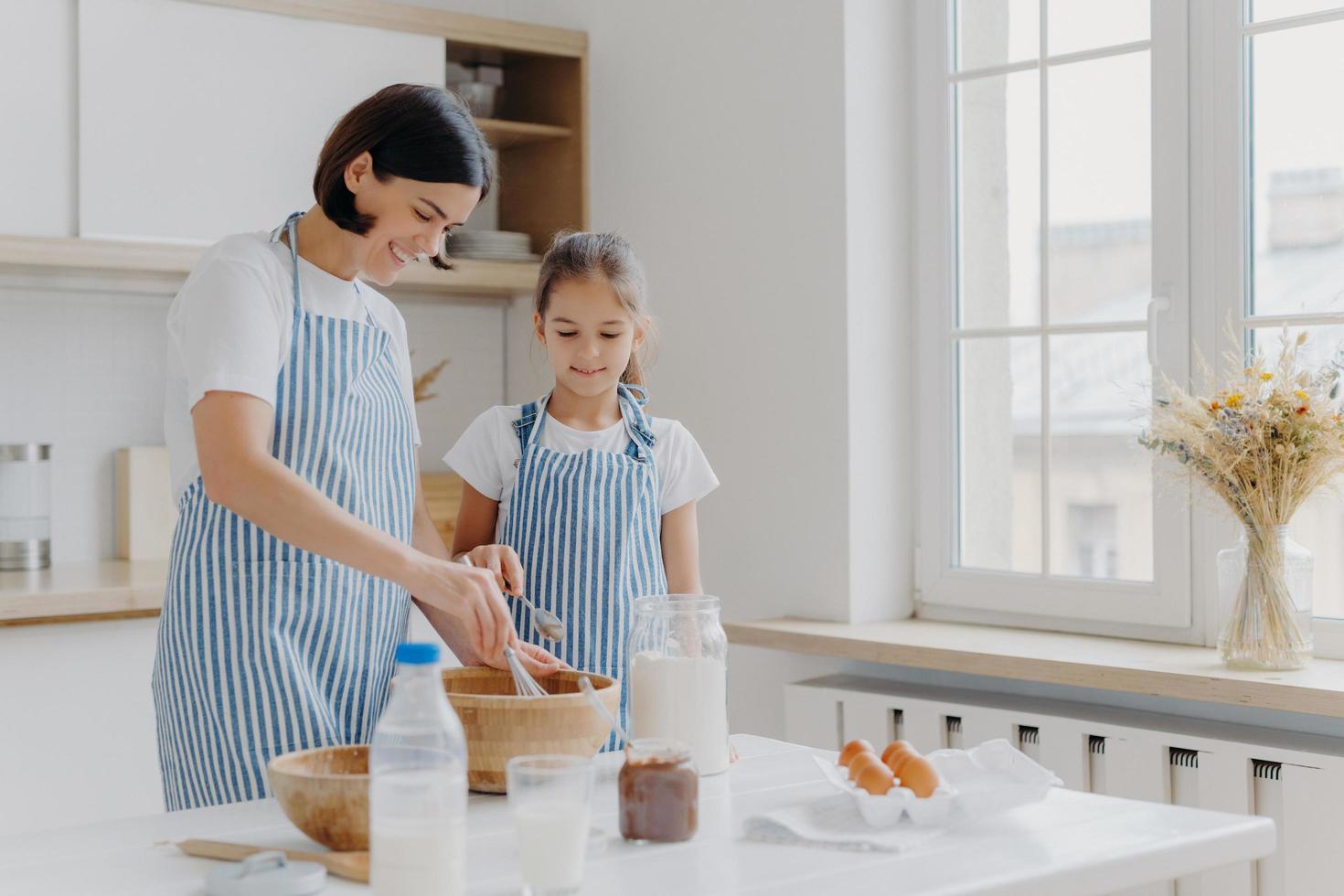 Horizontal shot of caring mother and daughter cook together, mom gives culinary lesson to small girl, use flour, eggs and milk to prepare tasty meal. Family time, baking and culinary concept photo