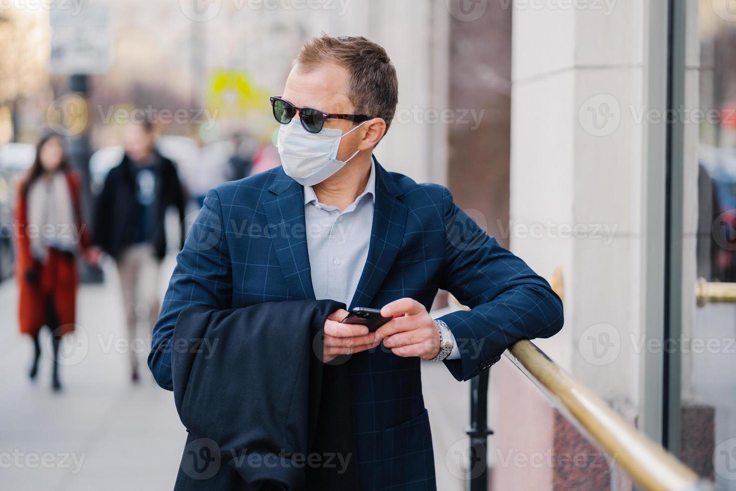 Prosperous businessman in formal wear poses at street, waits for someone, holds mobile phone and sends text messages, wears medical mask during coronavirus outbreak, few people walking outside photo