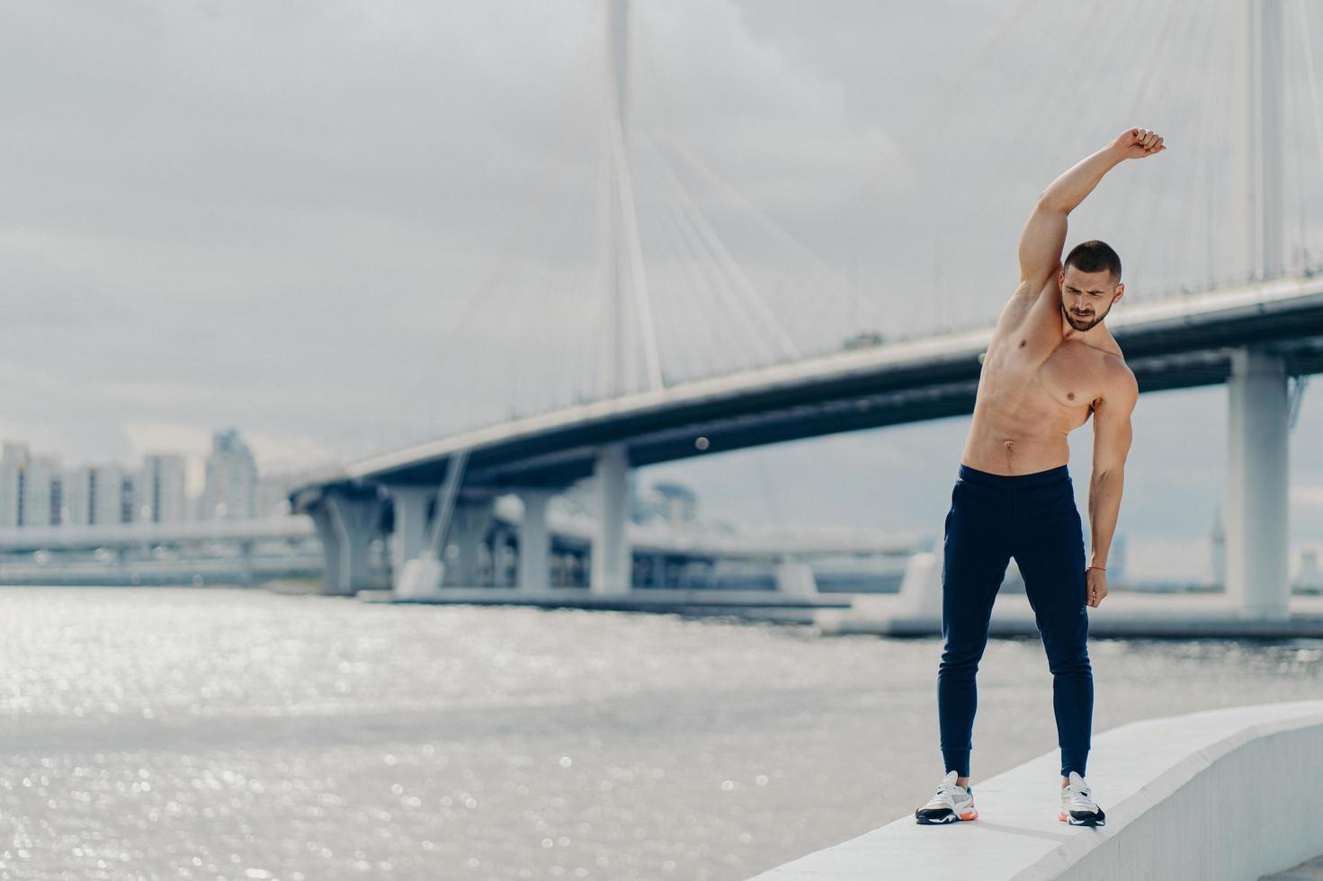 Full length shot of muscles active man does sport exercises outdoor poses near river bridge wears trousers and sneakers has beautiful male torso achieves best shape. Healthy lifestyle concept photo