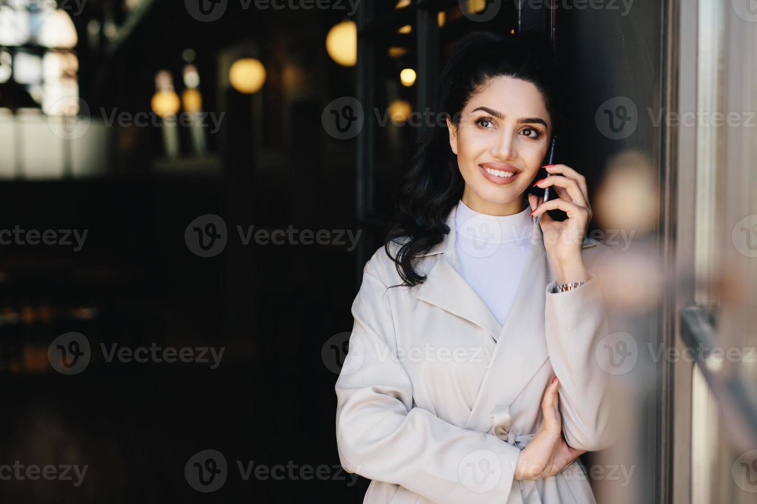 Elegant brunette businesswoman with dark eyes and full lips dressed in white clothes communicating over telephone having charming smile looking up with thoughtful expression. Technology concept photo