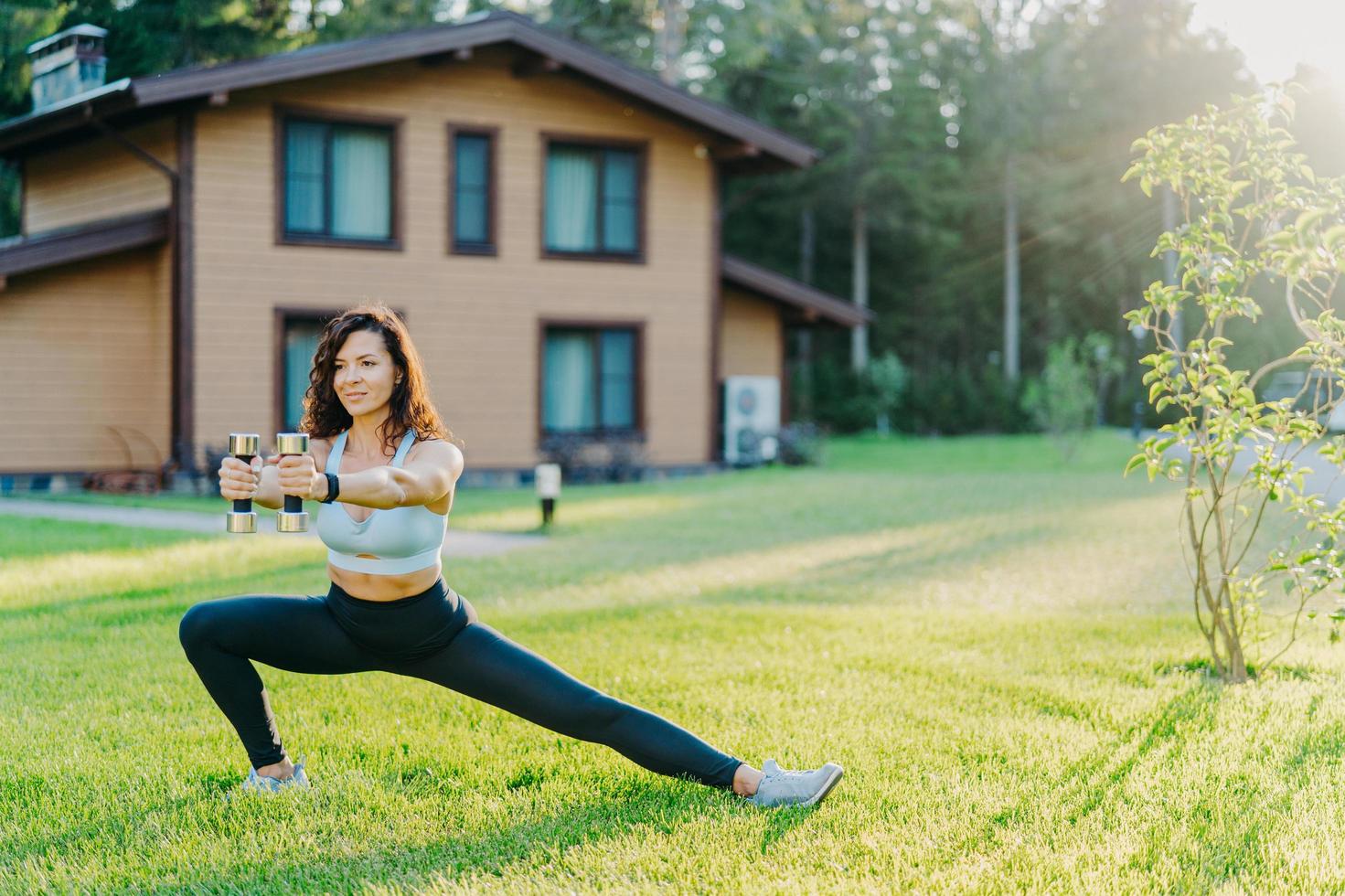 European sportswoman holds dumbbells and stretches legs, dressed in sport clothes, does physical exercises outdoor near house early in morning. Healthy lifestyle, weightlifting and fitness concept photo