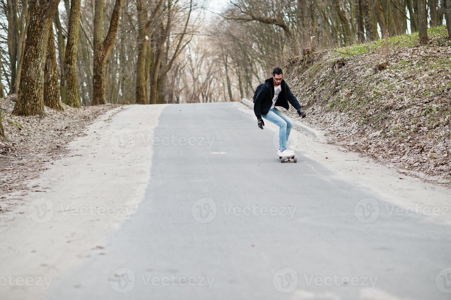 Street style arab man in eyeglasses with longboard longboarding down the road. photo