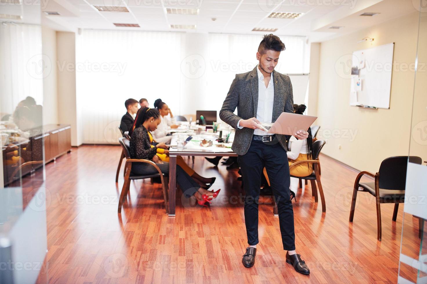 Face of handsome asian business man, holding laptop on the background of business peoples multiracial team meeting, sitting in office table. photo