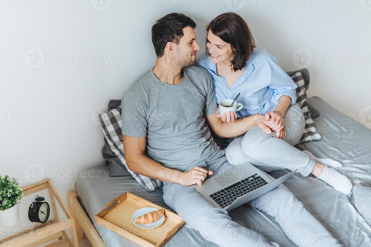 Affectionate female and male look at each other with great love, enjoy togetherness in bedroom, have good relationships, use laptop computer for entertainment, drink tea with croissant photo