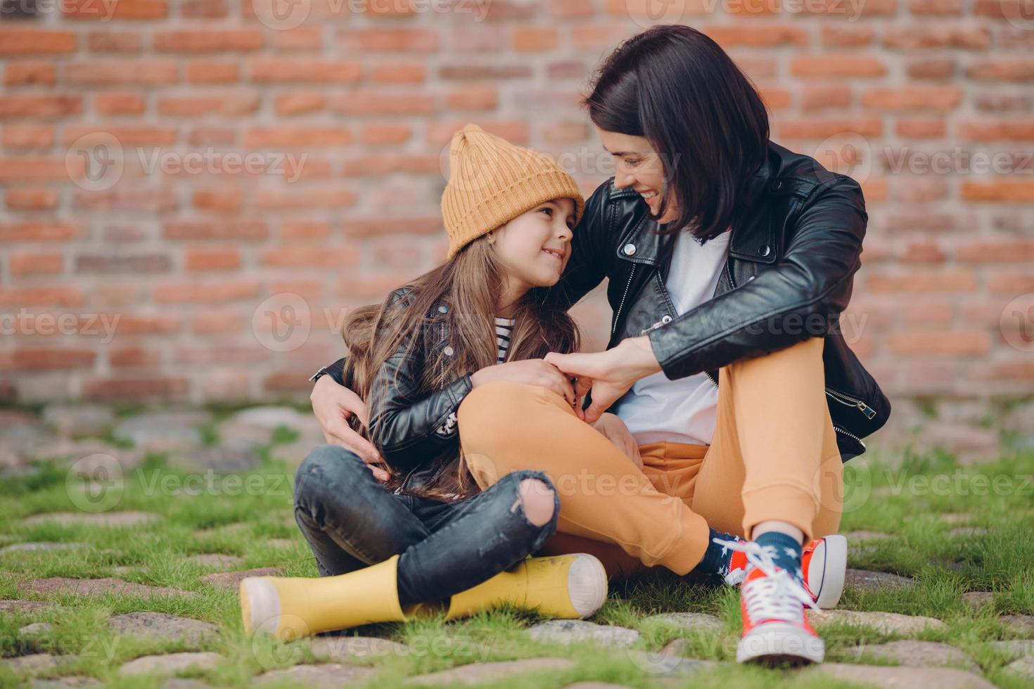 Lovely family of young mother and her small daughter embrace friendly, have good relationships, dressed in fashionable clothes, pose against brick wall outdoor. Parenthood and childhood concept photo