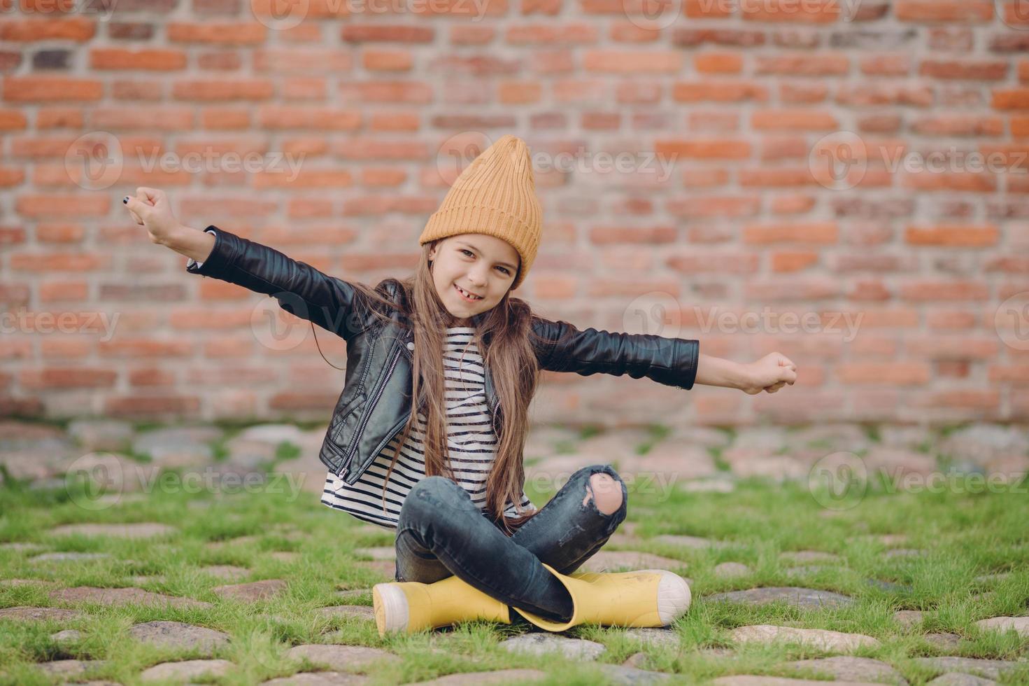 hermosa niña sentada en posición de loto, estira los brazos, usa sombrero amarillo, chaqueta de cuero y jeans amañados, posa sobre una pared de ladrillo al aire libre, tiene una expresión facial positiva. concepto de niños y diversión foto