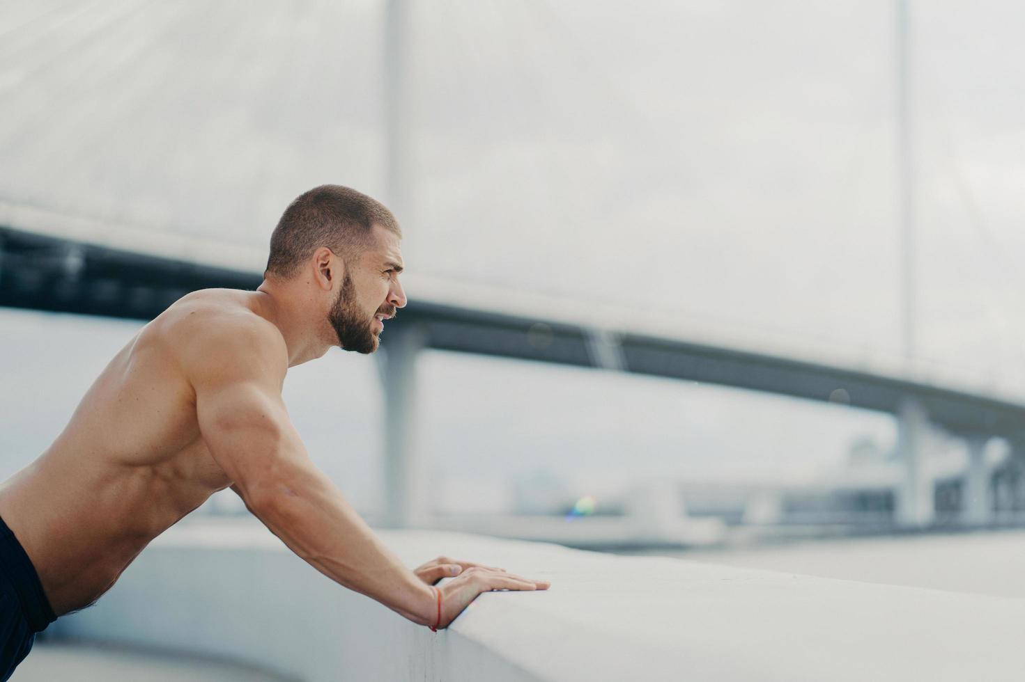 el deportista masculino motivado empuja hacia arriba las poses de ejercicio al aire libre en el puente concentrado en la distancia tiene entrenamiento matutino torso musculoso desnudo se calienta antes de trotar. concepto de estilo de vida saludable. foto