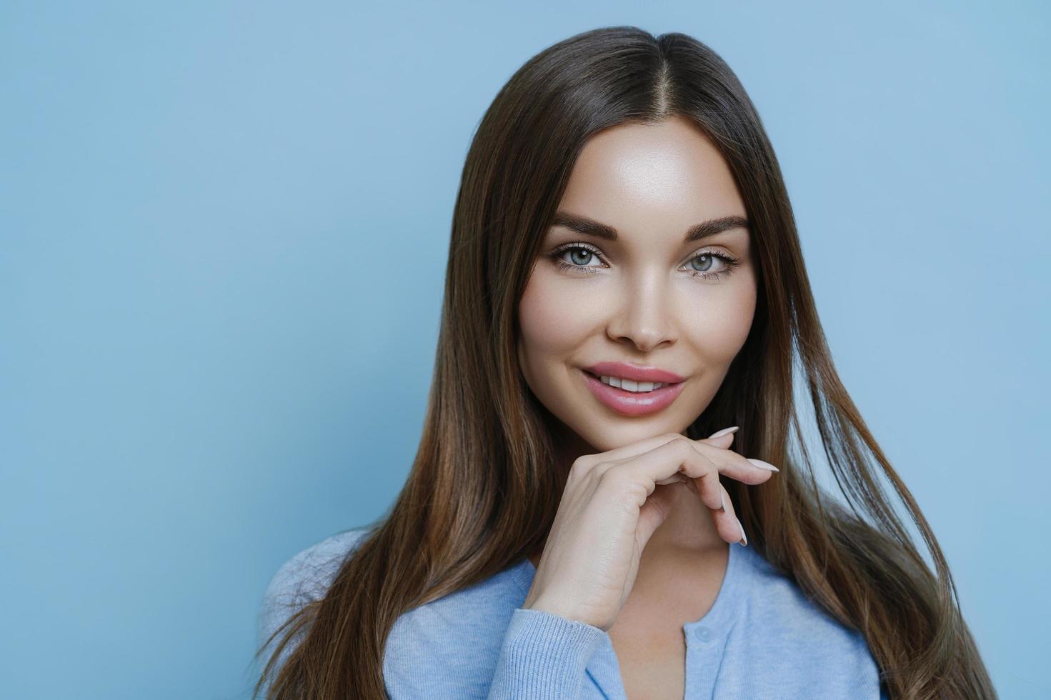 Isolated shot of attractive brunette woman with calm expression, healthy skin and makeup, keeps hand under chin, cares about complexion, looks beautiful, isolated on blue background, models in studio photo