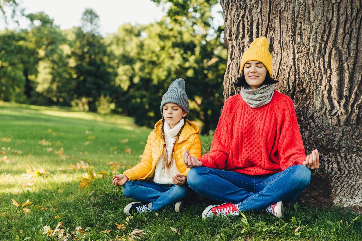 Relaxed carefree woman and little daughter sit in pose of lotus near tree in park, close eyes, try to concentrate, practice yoga together, dressed in warm clothes. Two female think about piece outdoor photo