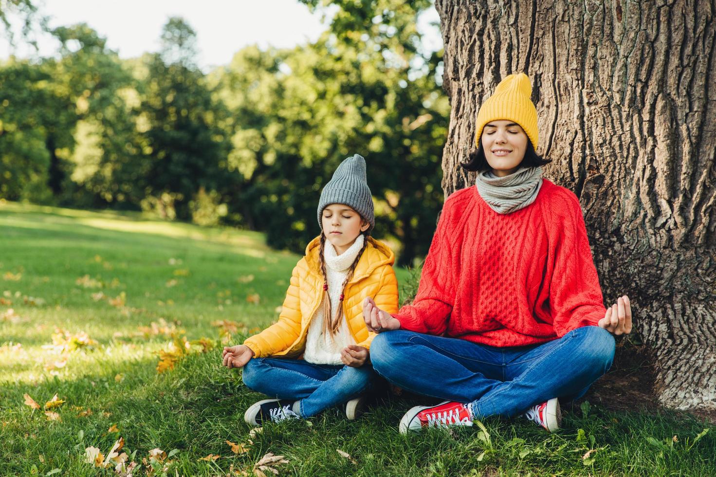 una mujer relajada y despreocupada y una hija pequeña se sientan en pose de loto cerca de un árbol en el parque, cierran los ojos, intentan concentrarse, practican yoga juntas, vestidas con ropa de abrigo. dos mujeres piensan en la pieza al aire libre foto