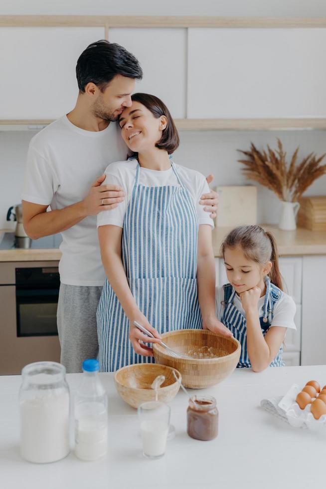 feliz y encantadora familia en la cocina de casa, el padre abraza a la madre con amor, la niña mira en el tazón, observa cómo mamá cocina y bate los ingredientes, usa huevos para hacer masa. ambiente domestico foto