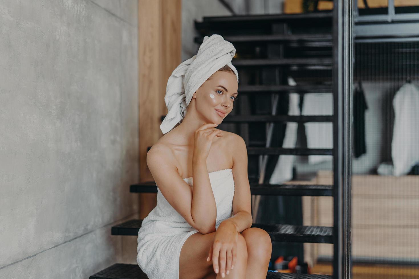 Thoughtful young European woman keeps hand under chin, sits on stairs and has thoughtful expression, has healthy skin, puts face cream, wrapped in bath towel, undergoes beauty treatments at home photo