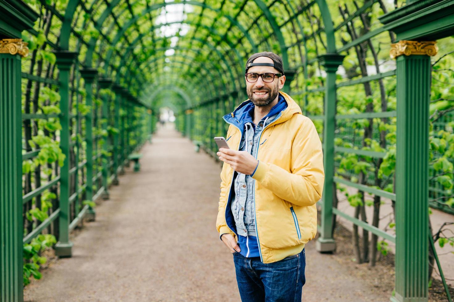 Young student male with beard having stylish clothes standing against green background using mobile phone for downloading video. Bearded hipster in yellow anorak resting outdoors typing messages photo
