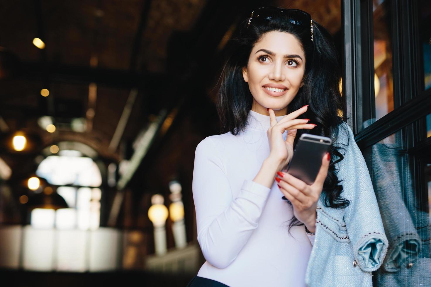 Portrait of thoughtful glamour woman with dark hair wearing sunglasses and white formal clothes surfing social networks using smartphone enjoying online communication posing against cafe interior photo