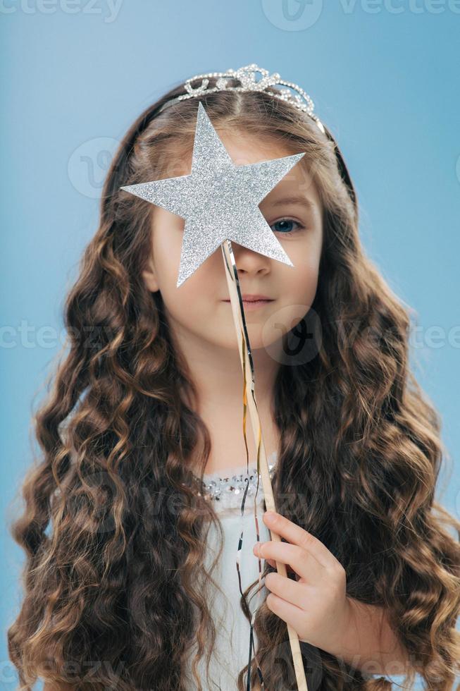 toma interior de una niña pequeña y guapa con el pelo largo y rizado, lleva corona, cubre la cara con una varita mágica en forma de estrella, aislada sobre un fondo azul claro. concepto infantil y mágico foto