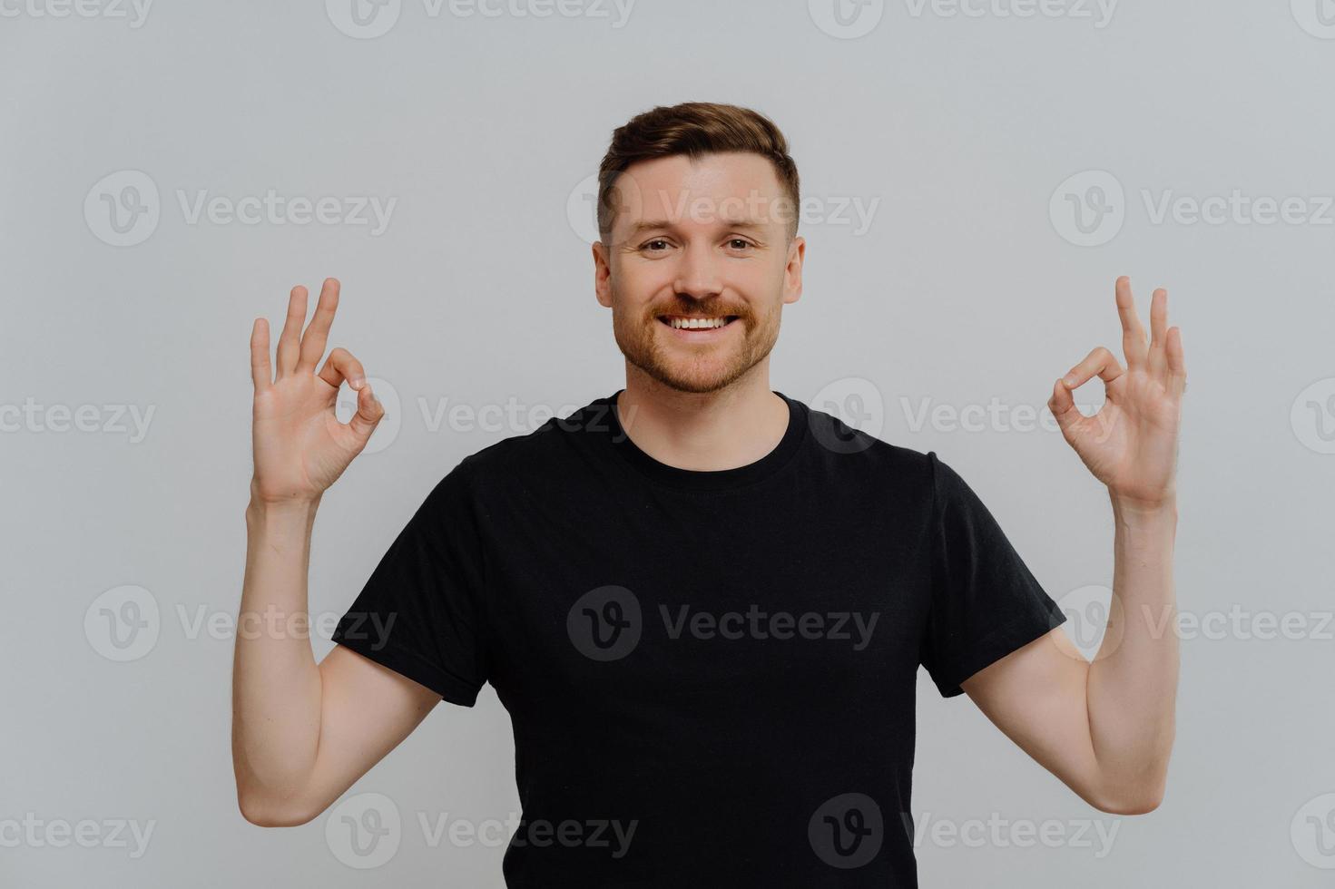 Young positive bearded man demonstrating agreement while showing okay gesture with both hands photo