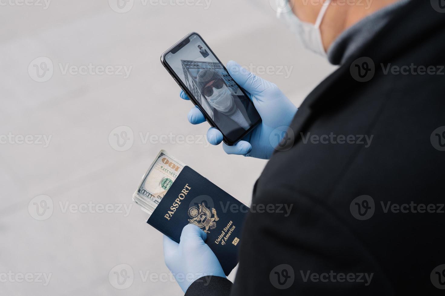 Shot of unrecognizable man follow rules of social distancing, makes video call, holds mobile phone, passport with money wears protective gloves walks outdoor at street. Avoid contact with other people photo