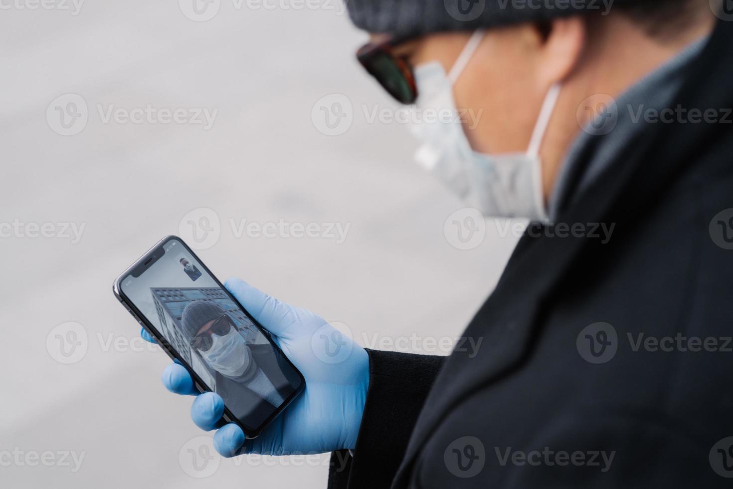 Cropped image of man has video call with best friend, keeps social distancing, being on self isolation during spreading infectious virus, wears protective mask and gloves, holds mobile phone in hand photo