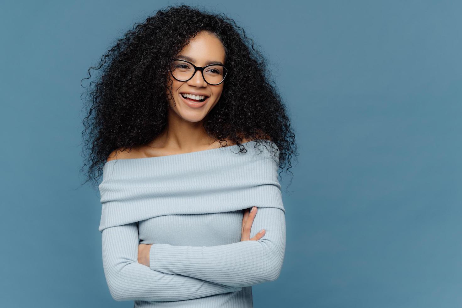 Cheerful female student has perfect mood, curly bushy hair, arms folded, looks somewhere with positive facial expression, wears blue sweater, stands indoor. People, feelings and emotions concept. photo