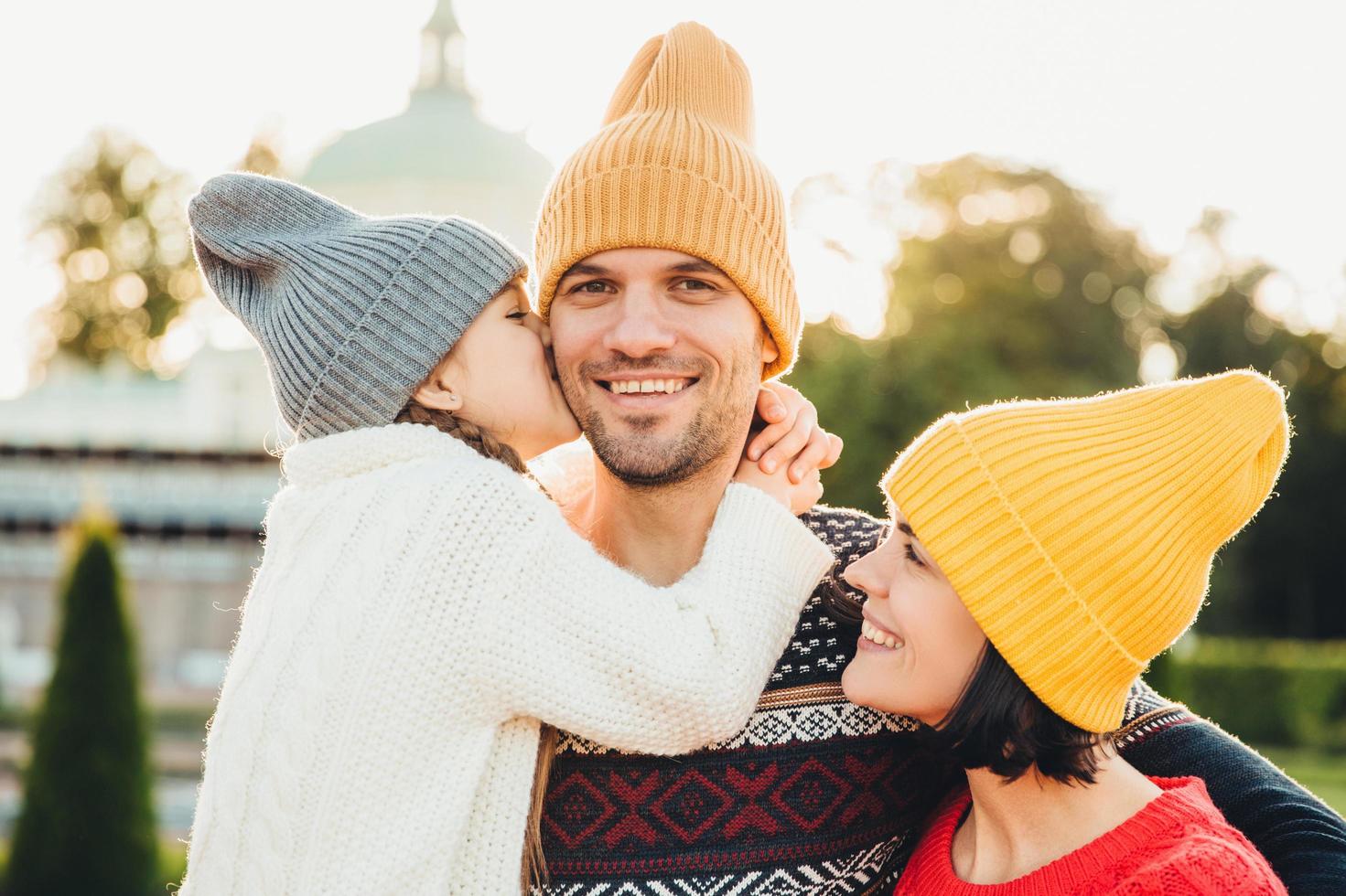 Pleasant looking little girl being thankful to her father for buying new bicycle, kisses him in cheek. Young family walk together outdoor, spend spare time in park, wear knitted fashionable hats photo