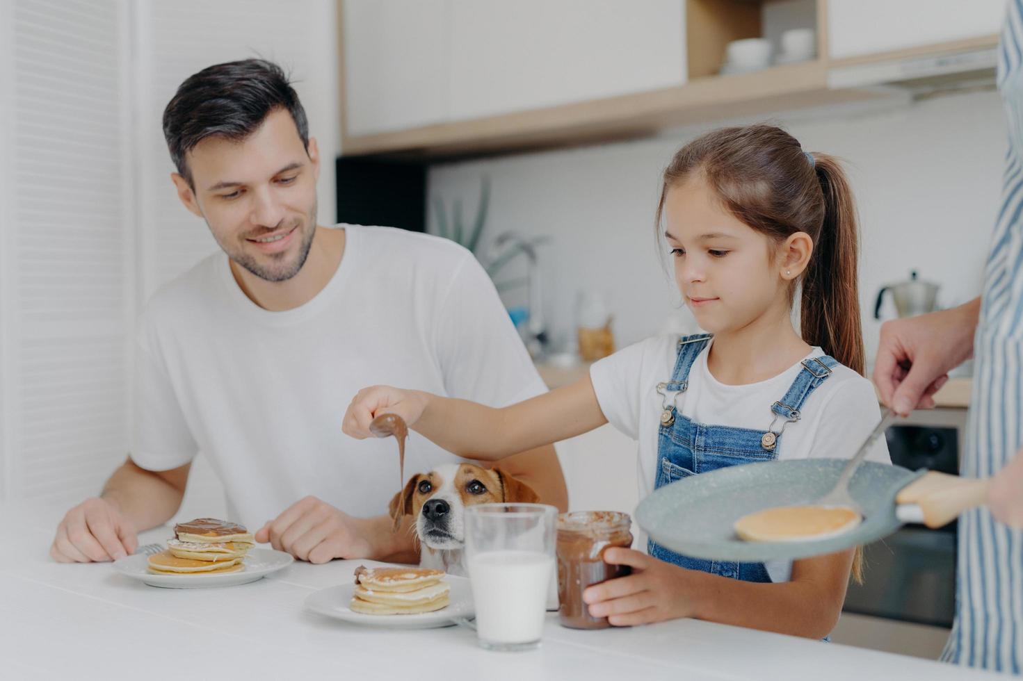 Photo of little girl in denim dungarees adds chocolate to pancakes, has breakfast together with dad and dog, likes how mother cooks. Family at kitchen have breakfast during weekend. Happy moment