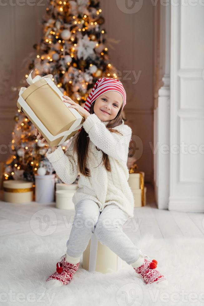 Vertical portrait of delightful small female child in white clothes and warm socks holds big present box, recieves gift from parents on New Year, poses against decorated Christmas tree photo